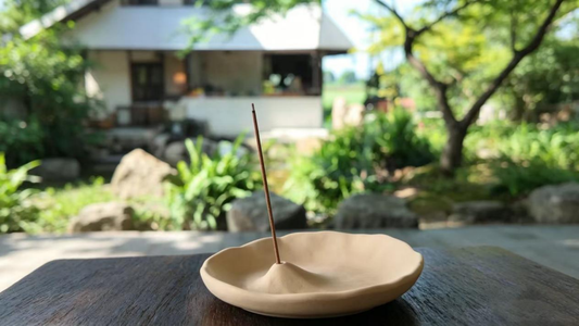 Minimalist incense stick burning in a beige ceramic holder, set on a dark wooden table in a peaceful Japanese garden with lush greenery and a traditional house in the background, creating a calm, meditative atmosphere.